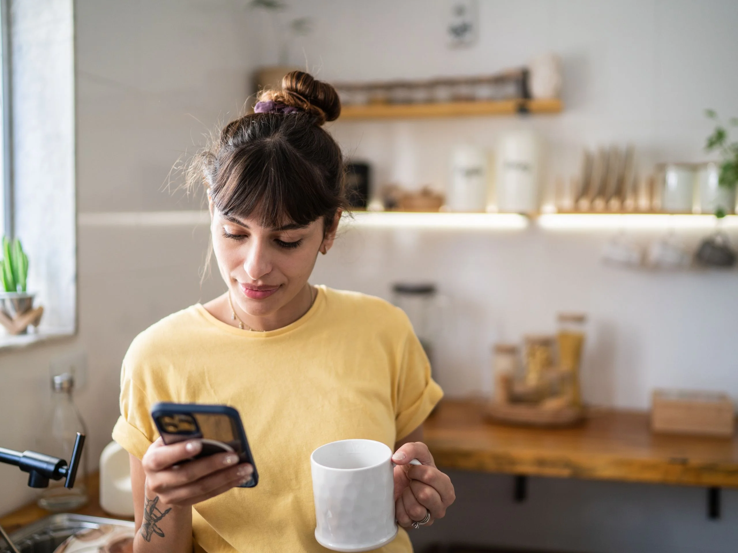 Mujer joven en la cocina con una taza de café y mirando el móvil