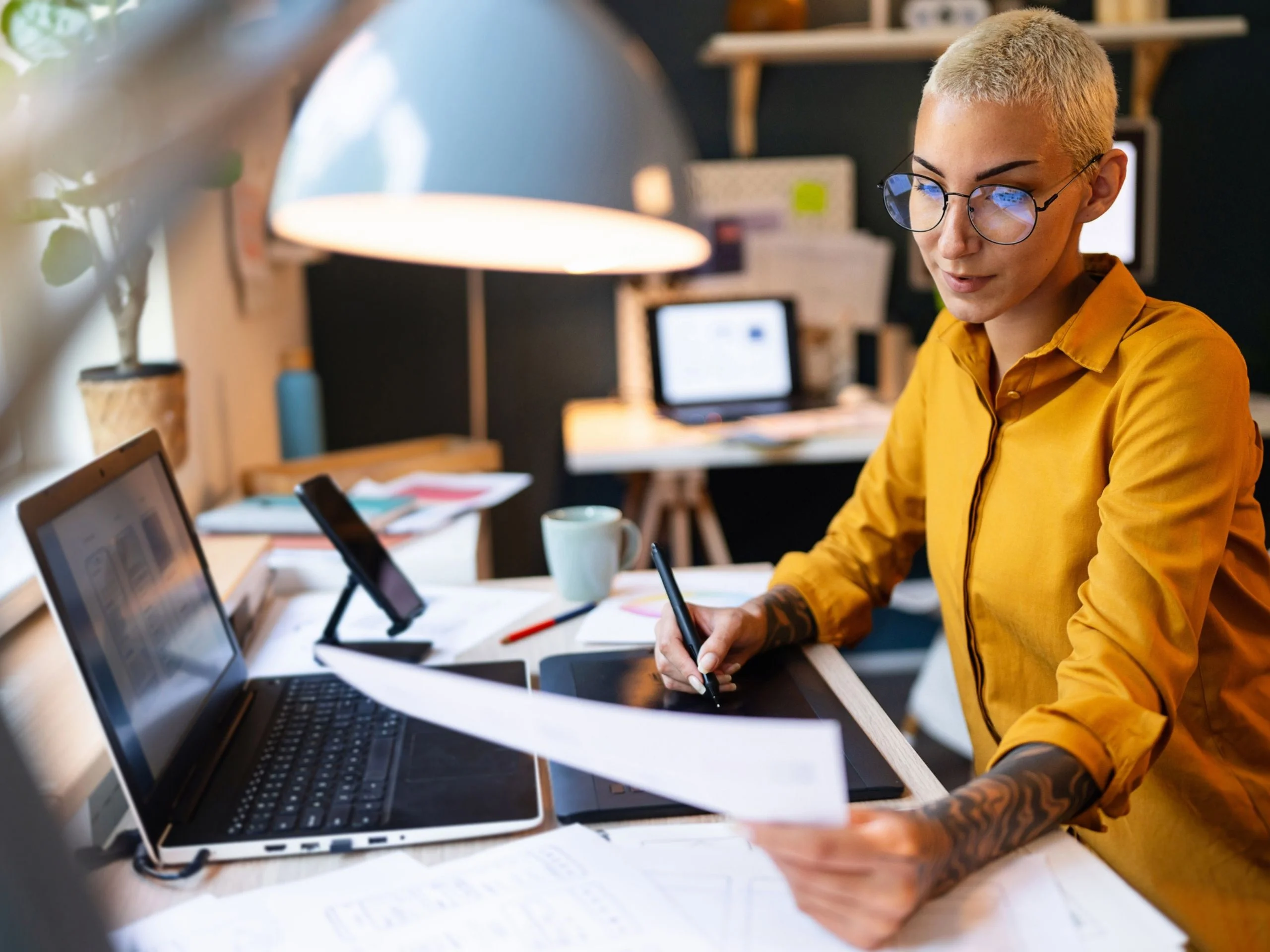 Mujer con gafas y tatuajes revisando documento y tecleando en portátil