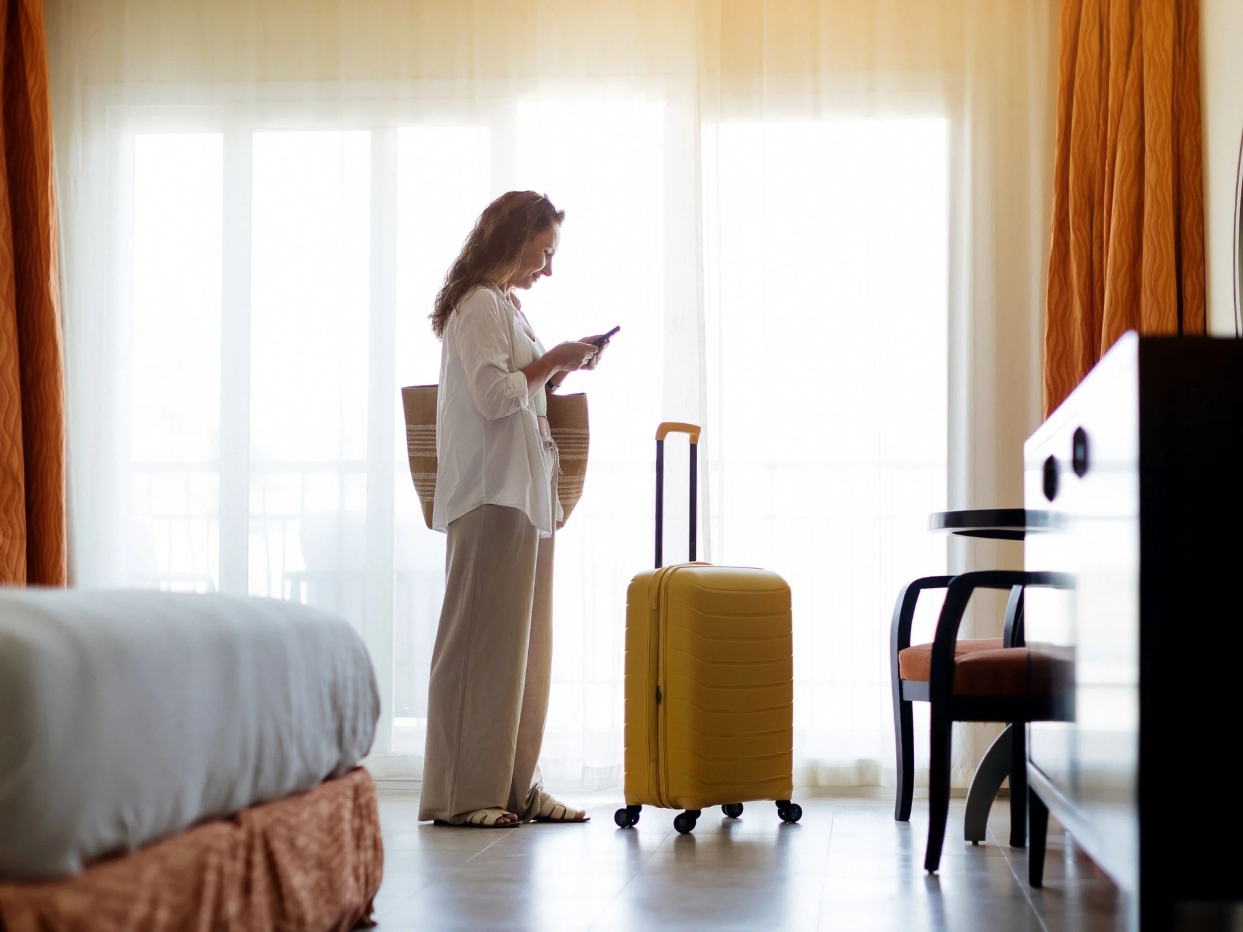 Mujer de pie en la habitación del hotel con una maleta amarilla, mirando su teléfono