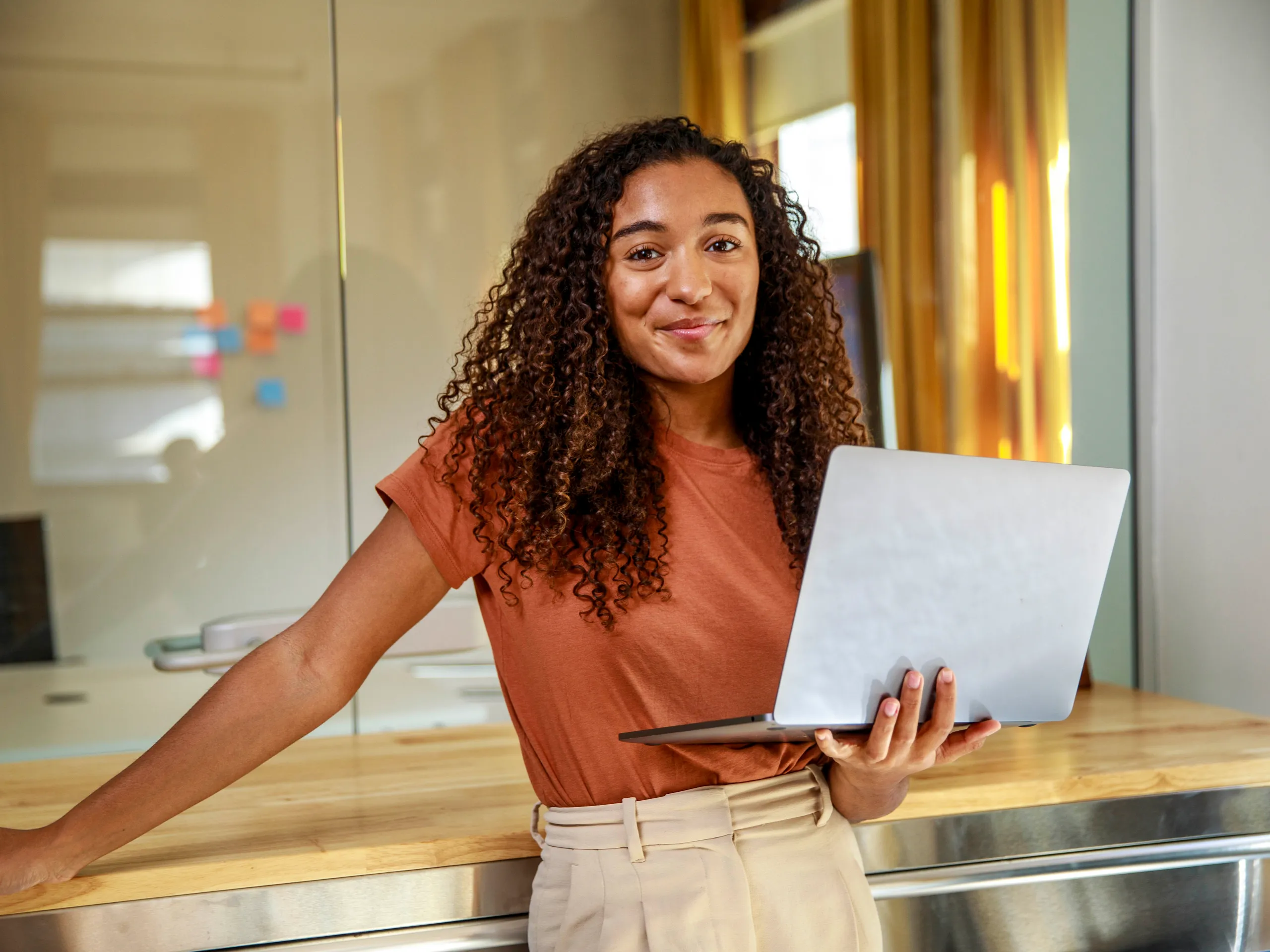 Smiling woman with curly hair holding a laptop in a modern office.