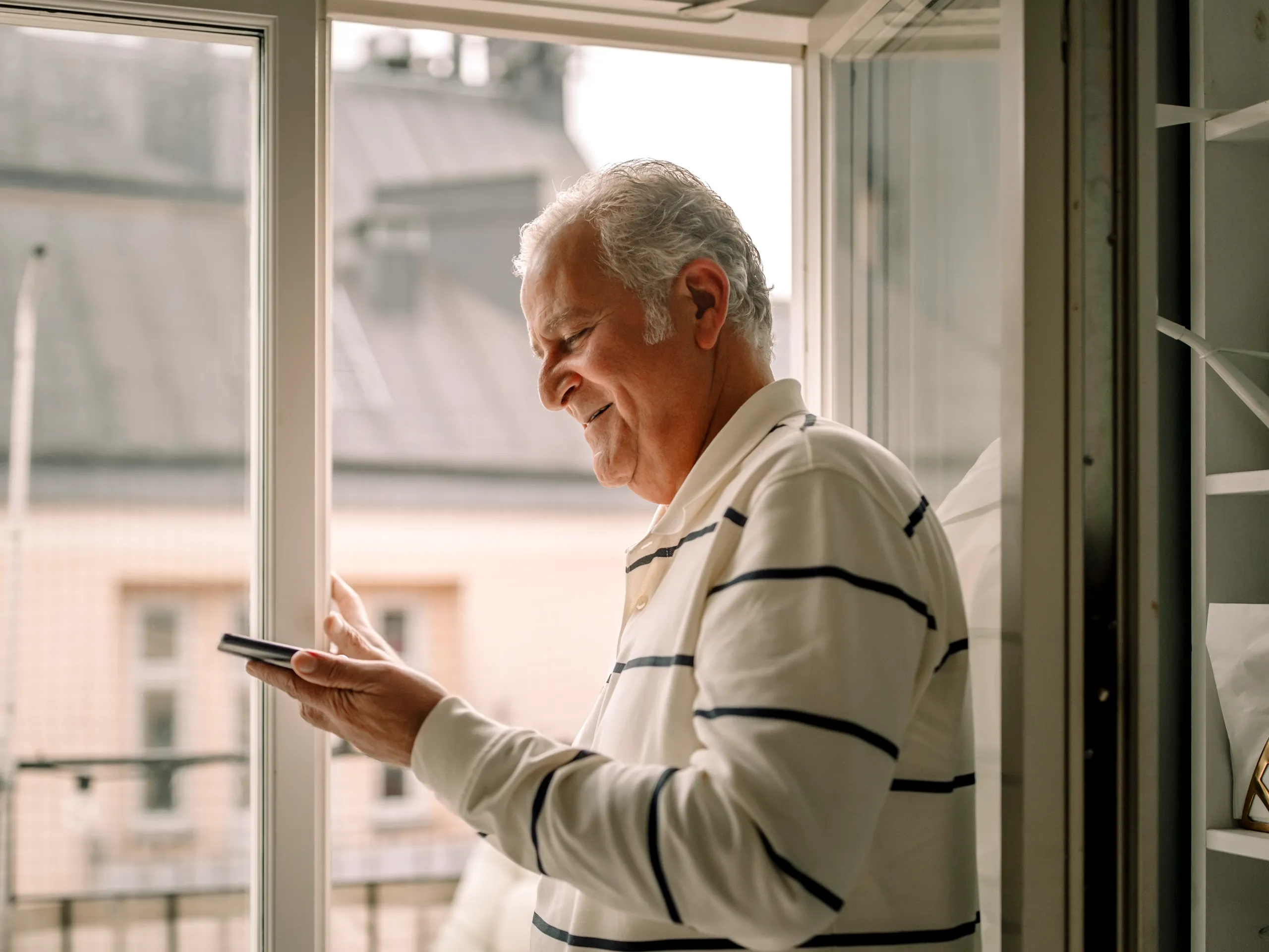 Older man using a smartphone near a sunny window.