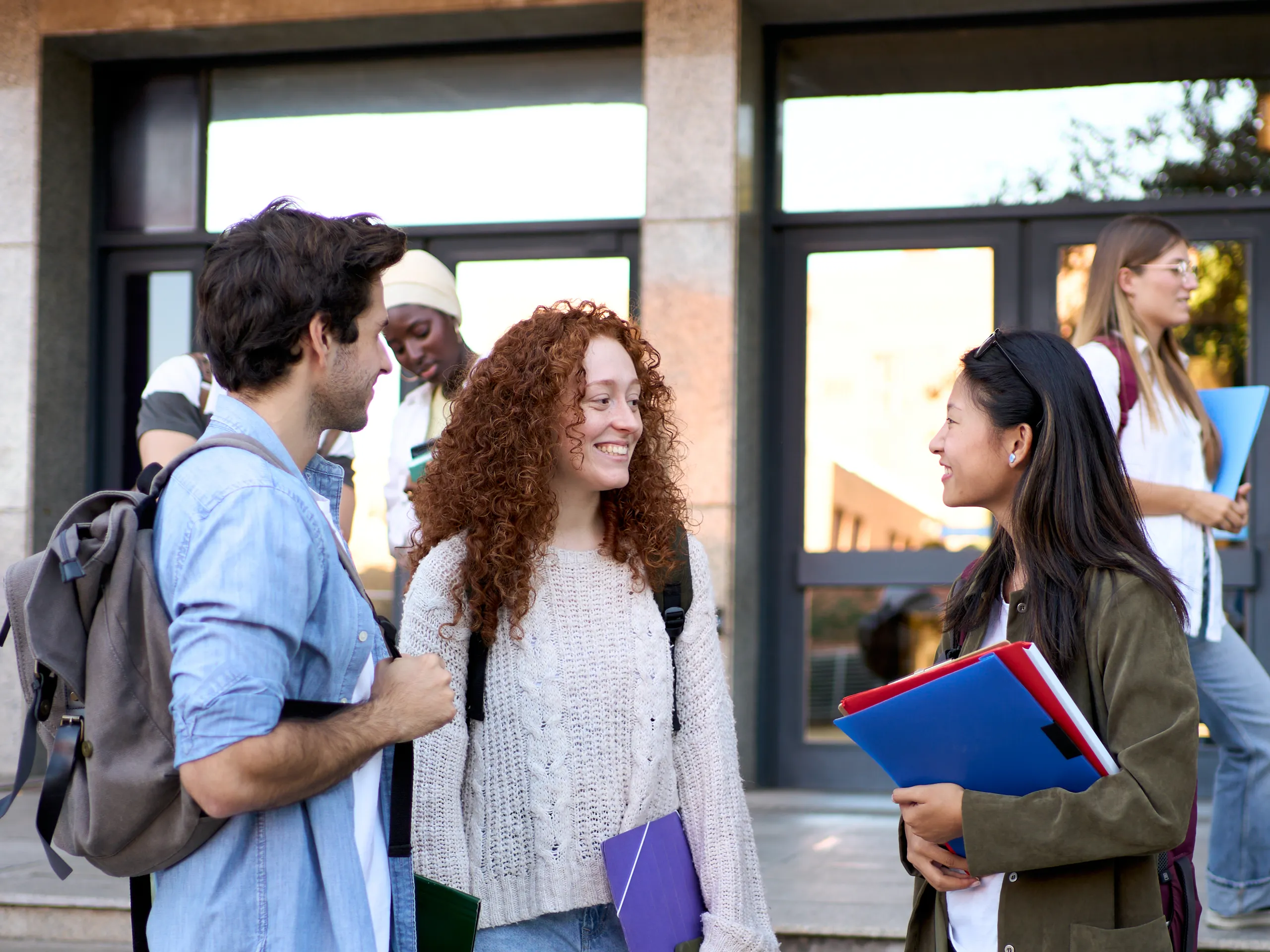 Tres jóvenes sonríen y hablan frente a un colegio.