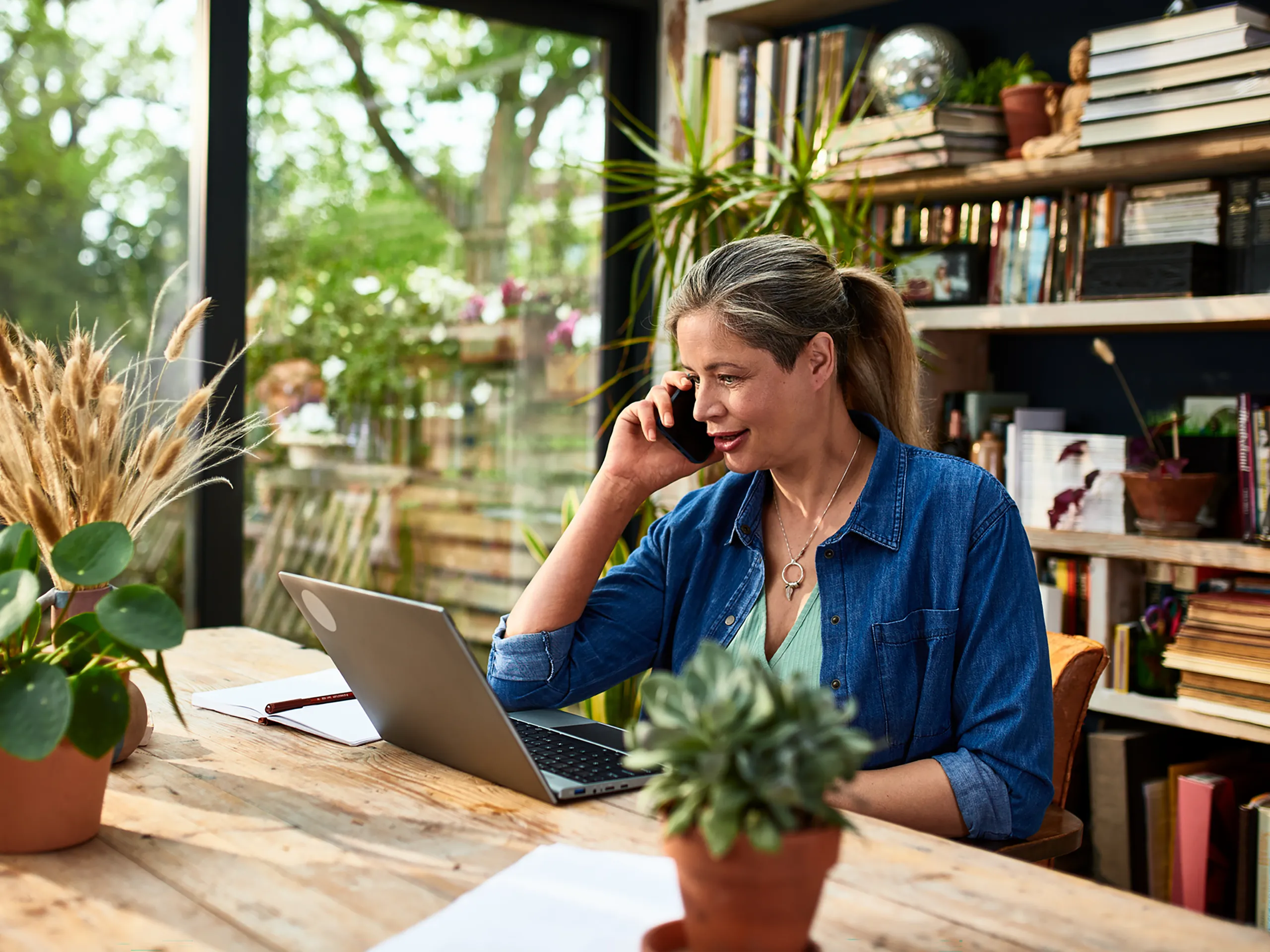 Una mujer habla por teléfono mientras trabaja con su computadora portátil en una habitación luminosa con plantas.