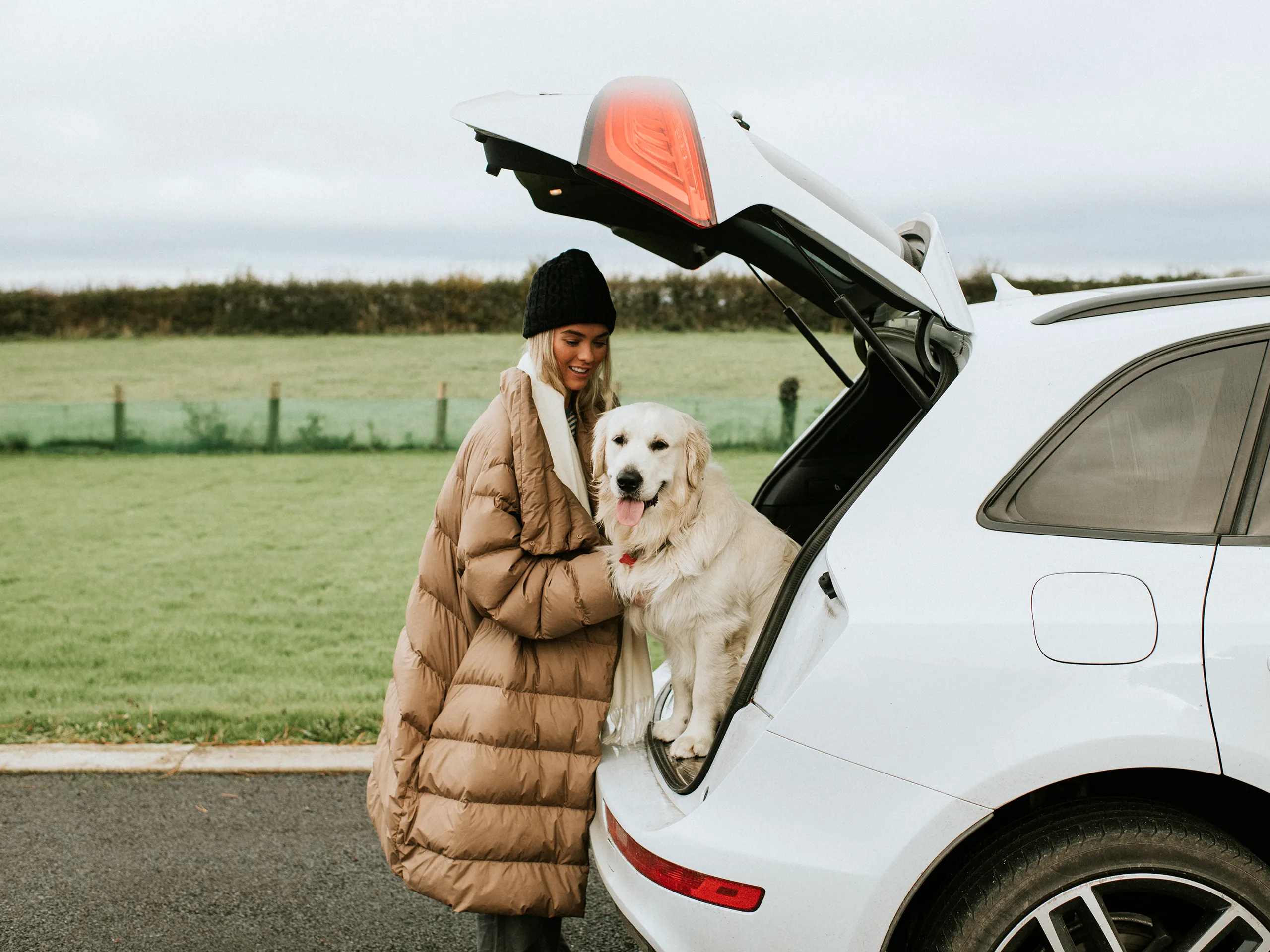 Woman with a golden retriever stands next to a car with an open trunk.