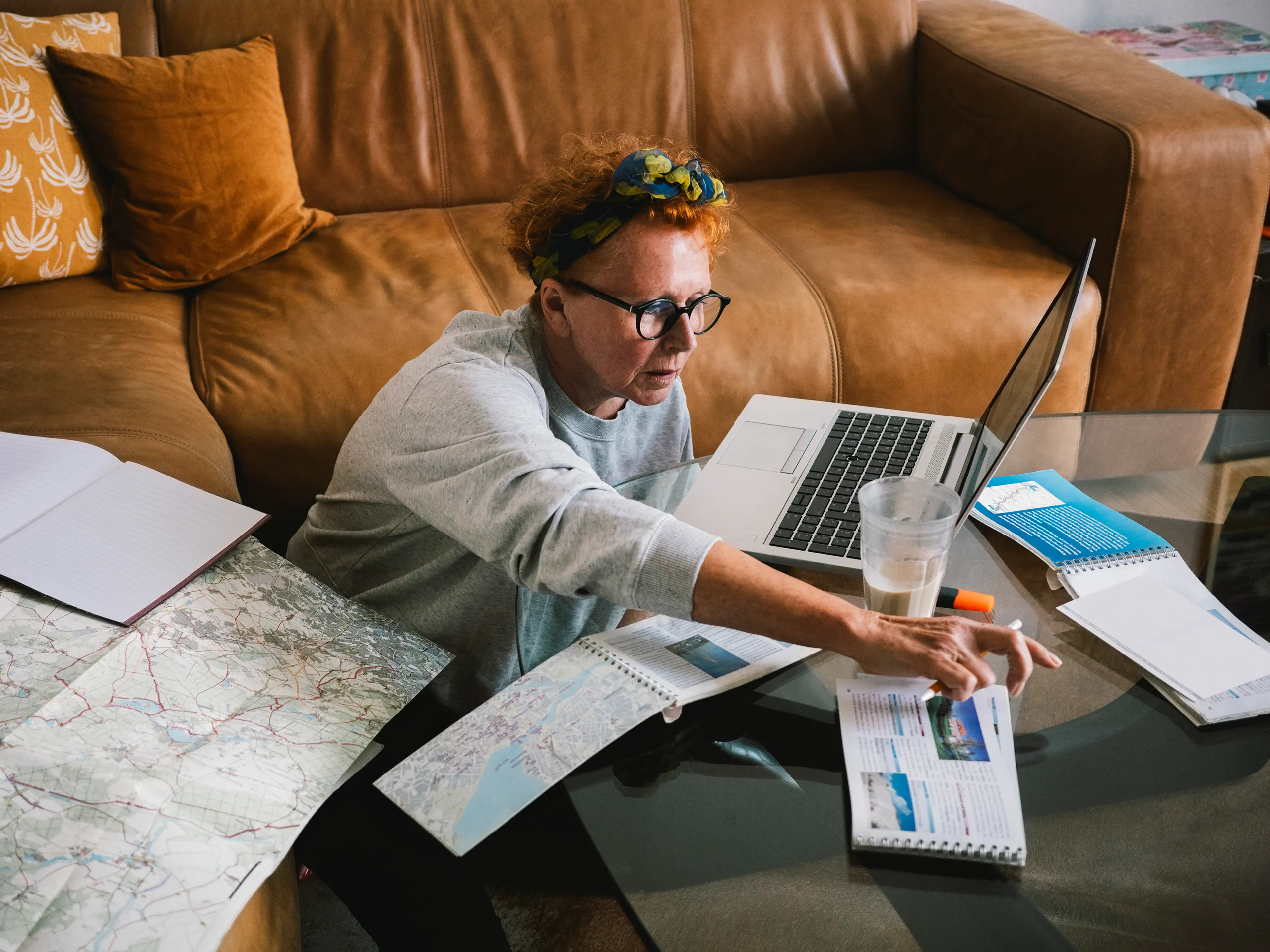 Mujer rodeada de mapas y una computadora portátil planeando un viaje en casa.