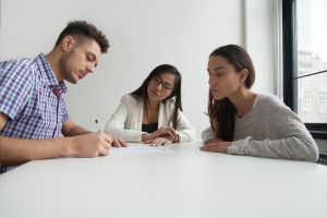 Hombre blanco firmando un documento en una mesa. Frente a él, mujer blanca mirando atentamente. Entre los dos, mujer asiática revisando la acción