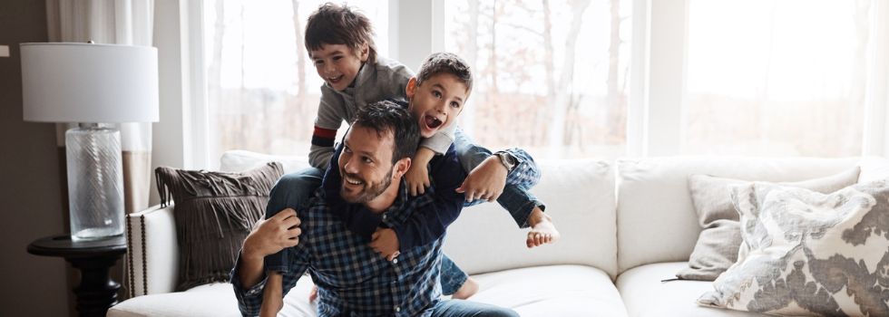 Padre con dos hijos subidos encima, jugando, en el sofá de casa