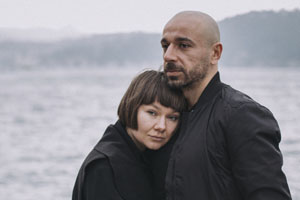 Pareja de hombre y mujer blancos vestidos de negro y abrazados con el mar de fondo