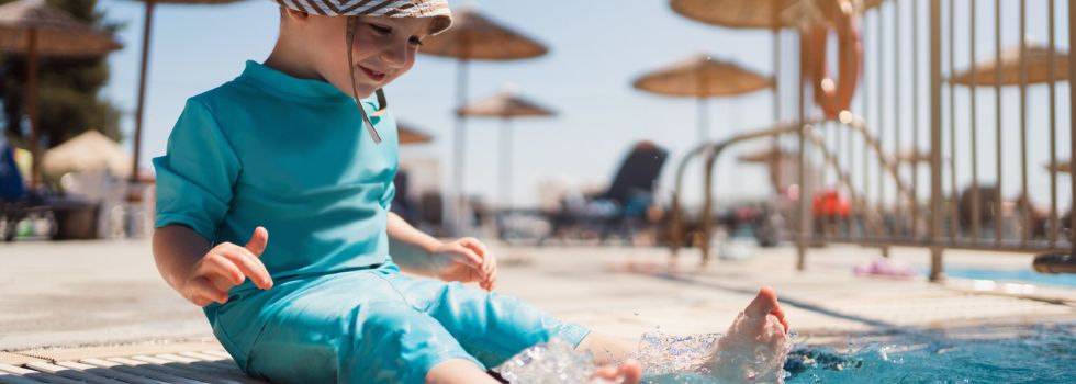 Niño blanco con bañador de cuerpo entero y gorro en el borde de la piscina, chapoteando con los pies