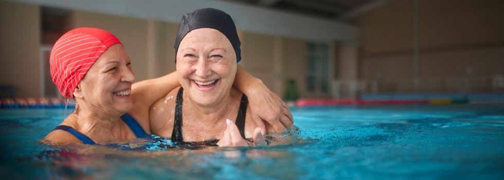 dos mujeres blancas mayores con gorro de natación en una piscina abrazadas y riendo 