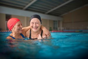 dos mujeres blancas mayores con gorro de natación en una piscina abrazadas y riendo 