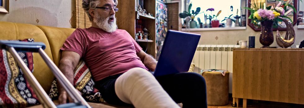 Hombre blanco con barba en el sofá mirando el portátil y con una escayola en la pierna