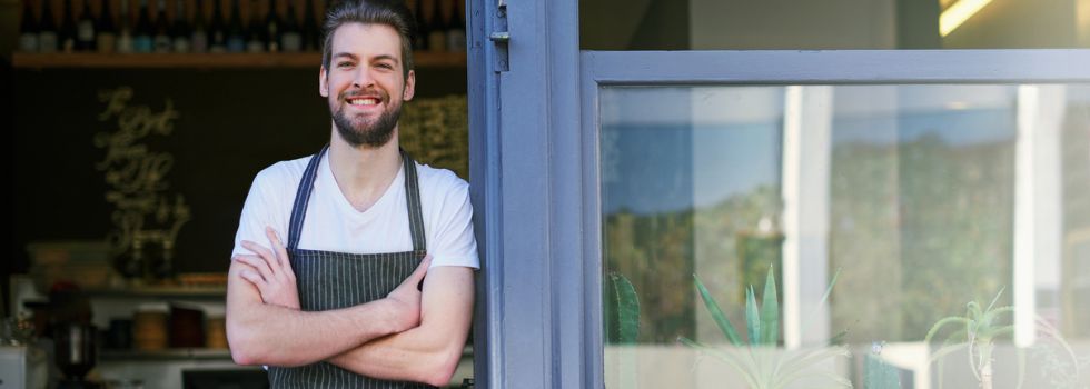 Hombre blanco frente a su cafetería, apoyado en la puerta y sonriendo
