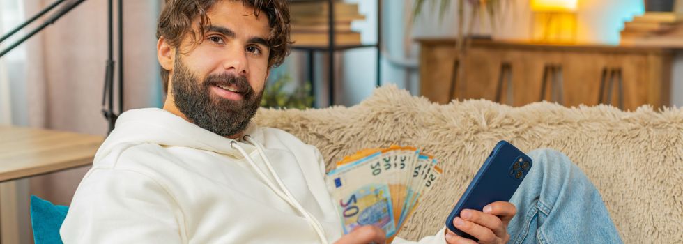 Hombe indio en casa sentado en el sofá con un teléfono en una mano y en la otra un fajo de billetes de euro