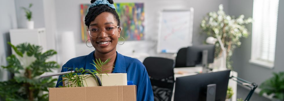 Mujer negra con gafas feliz deja la oficina con una caja de cartón con sus cosas 