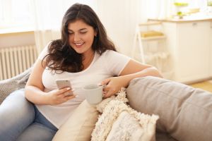 Mujer morena con sobrepeso mirando su móvil en el sofá mientras bebe un café