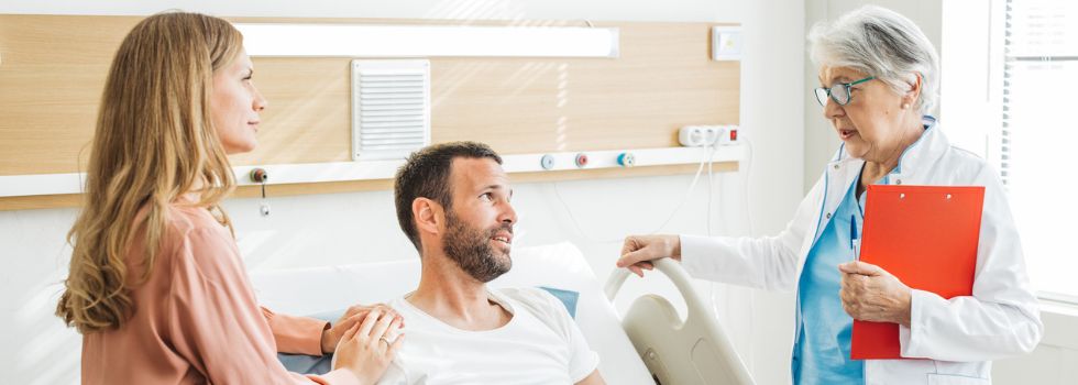 Hombre en cama de hospital con mujer a su lado y médico hablando a los dos