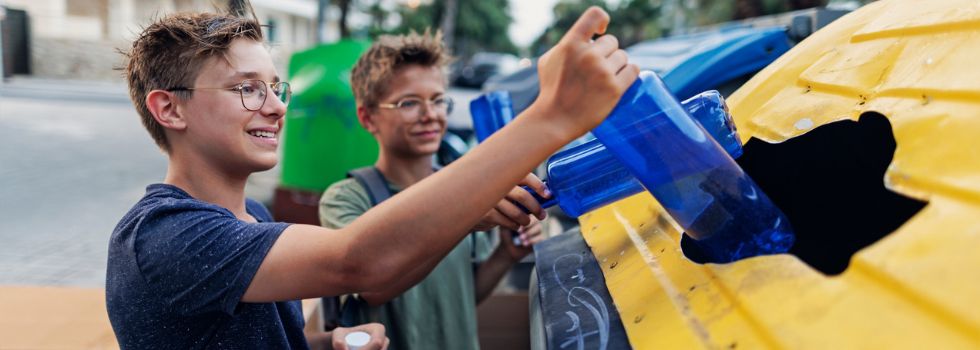 Dos niños blancos y con gafas tirando botellas de plástico al contenedor amarillo de reciclaje