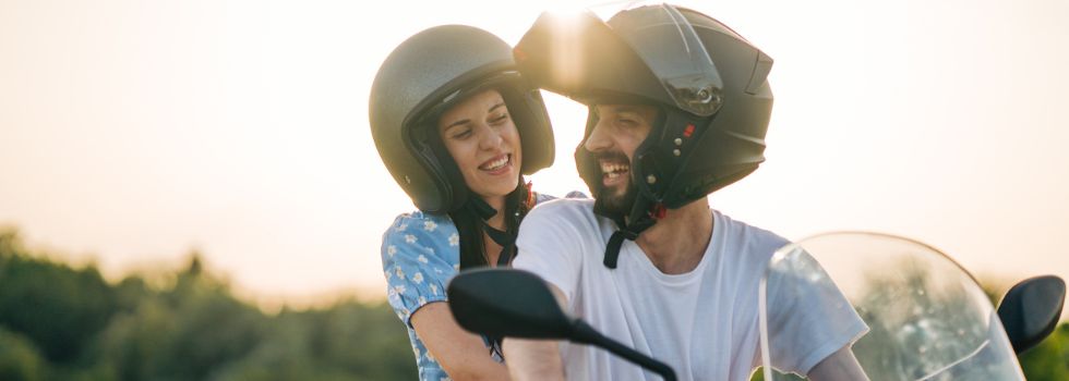 Hombre y mujer jóvenes y blancos montados en una moto, sonriéndose entre ellos