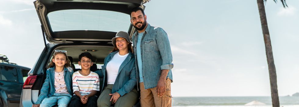 Familia negra sentados en la parte de atrás de un coche, con el maletero abierto. Detrás, una playa