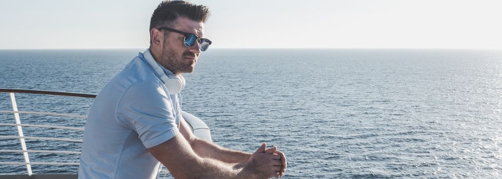 Hombre blanco con gafas de sol apoyado en la cubierta de un barco mirando al horizonte