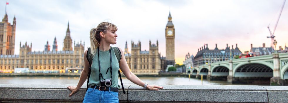 Mujer blanca con cámara de fotos apoyada en muro a la orilla del Támesis. El Parlamento inglés y el Big Ben de fondo.