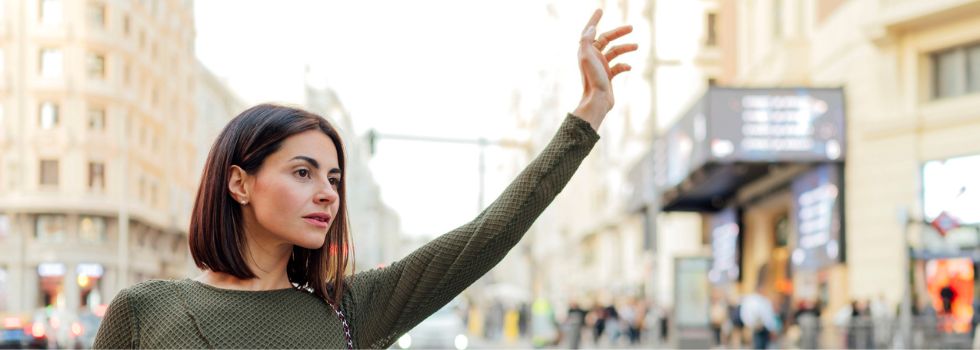 Mujer blanca con el brazo en alto, parando un taxi en la calle